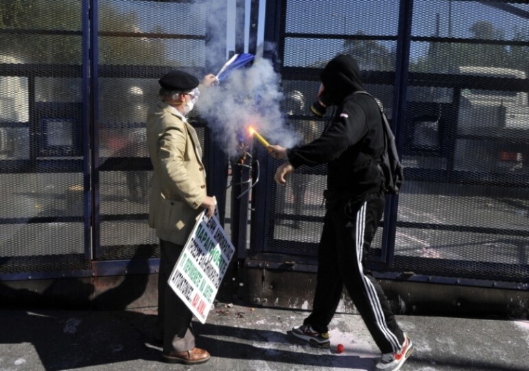 Ad-Atene-un-anziano-da-fuoco-alla-bandiera-insieme-ad-un-manifestante-incappucciato