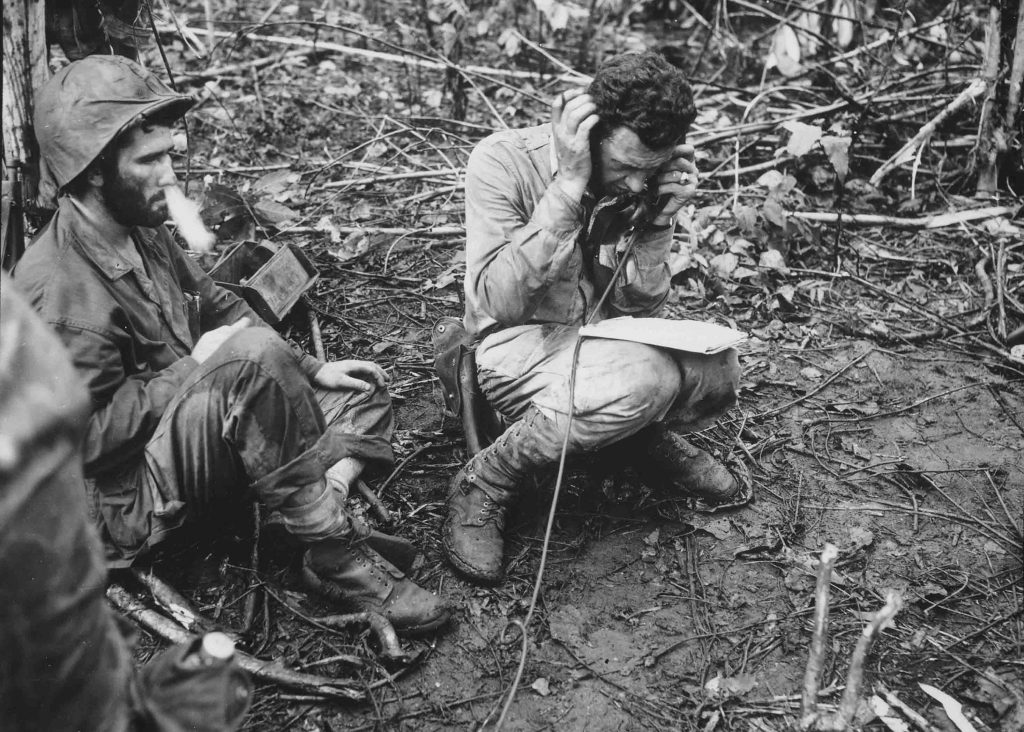 Il tenente colonnello John Weber, comandante di un battaglione di marines a Cape Gloucester, seduto sul suo elmetto, riceve un rapporto da uno dei suoi comandanti di compagnia. Il soldato semplice di prima classe Vincent Miley, guardando davanti a sé, fa uscire il fumo di una sigaretta dal naso. Gennaio 1944, Cape Gloucester, Papua Nuova Guinea Autore sconosciuto o non fornito © courtesy Defense Dept. photo (Marine Corps) / Library of Congress