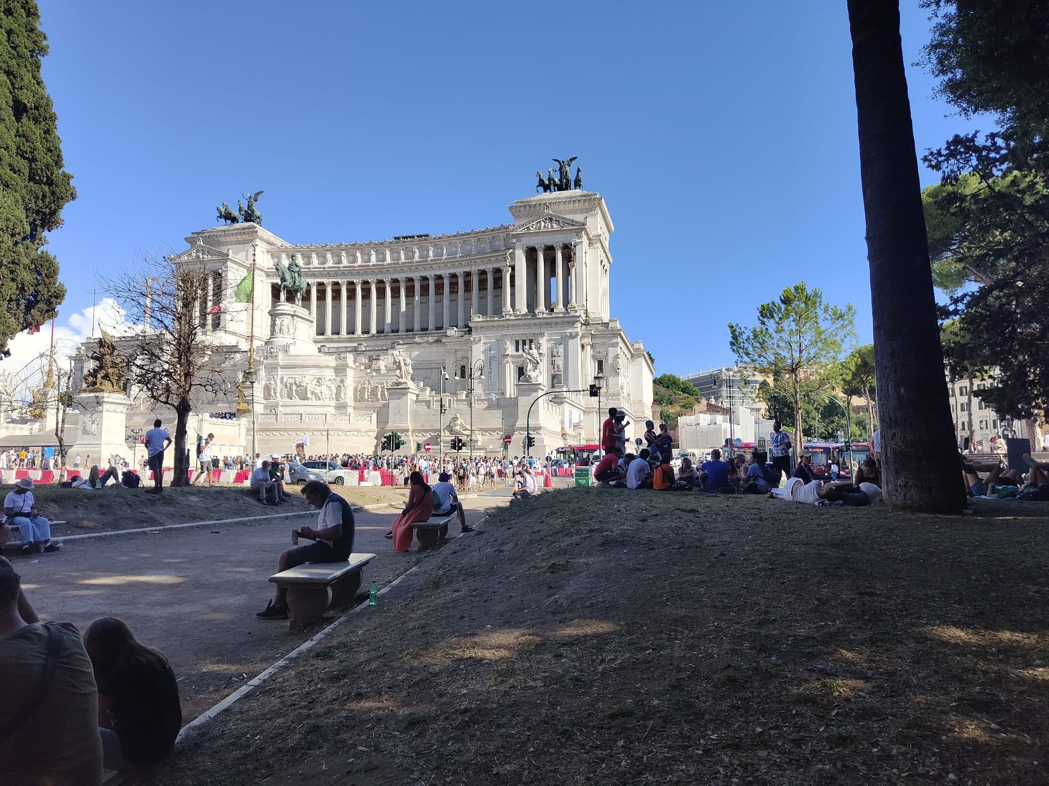 Gruppi di pellegrini (e non solo) trovano riparo dal sole sotto agli alberi davanti all’Altare della Patria nel caldo pomeriggio di luglio.